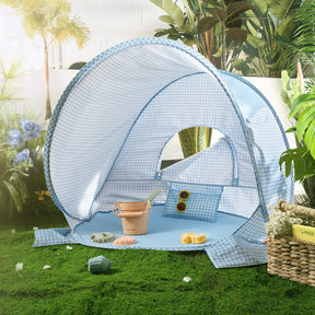 The Tiny Land® One-Stop Beach Toy & Tent Set in light blue gingham sits on grass, with a pillow, yellow sunglasses, beige bucket, and sand molds inside. Flowers and plants surround the UV-protective tent as sunlight shines in.