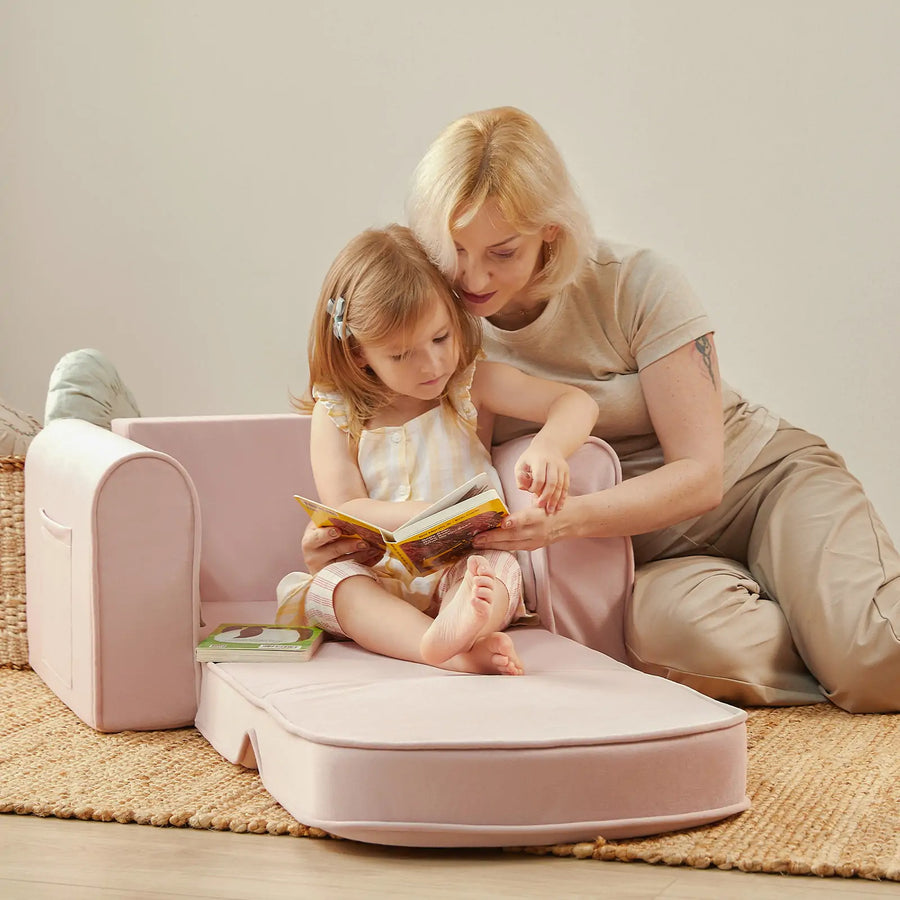 A woman sits on the floor with a young girl on the Tiny Land® GentleSnug Kid Chair, reading a book together in a cozy, softly lit room. This 2-in-1 sofa bed by Tiny Land, crafted from high-density foam, adds comfort and versatility to their special moment.|Pink