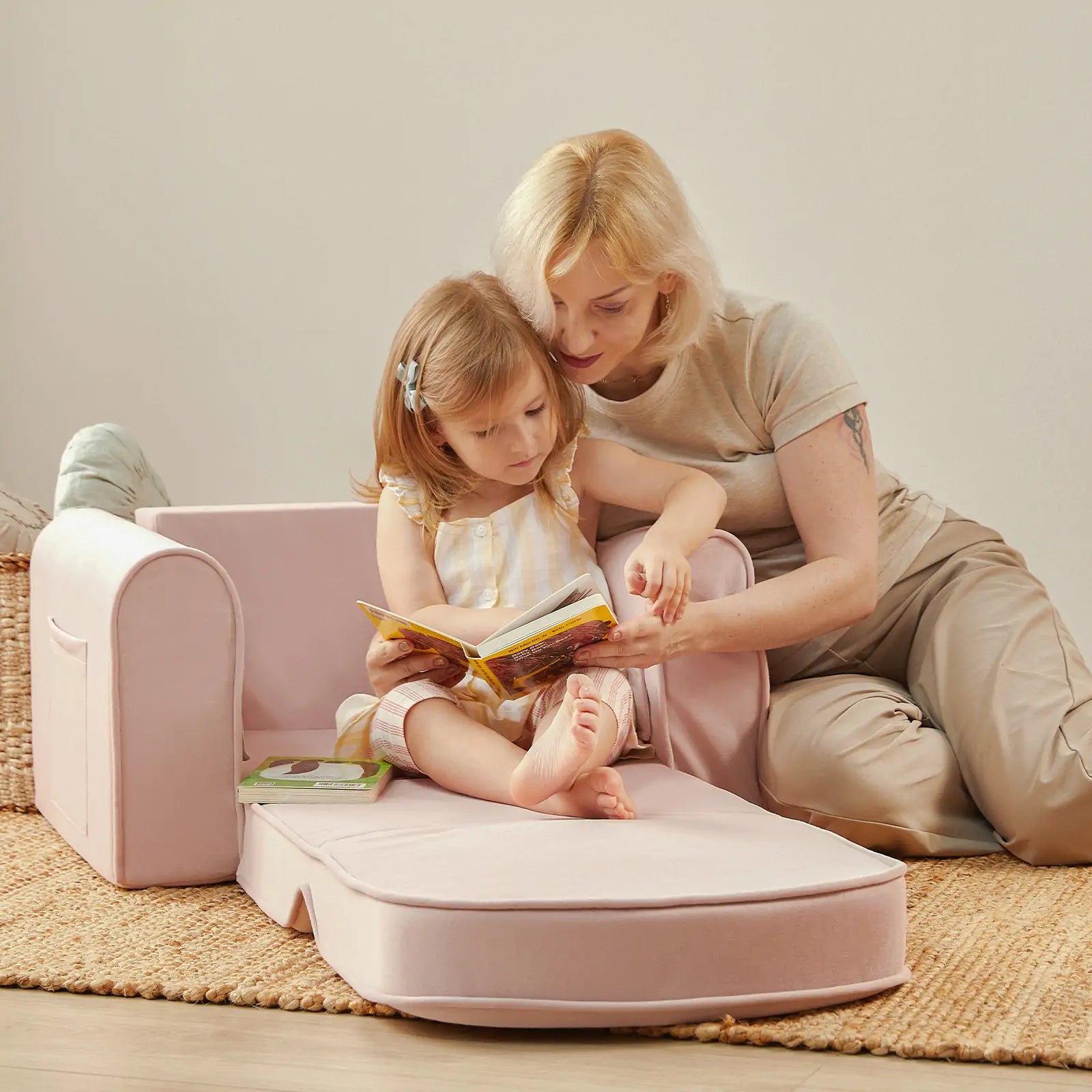 A woman sits on the floor with a young girl on the Tiny Land® GentleSnug Kid Chair, reading a book together in a cozy, softly lit room. This 2-in-1 sofa bed by Tiny Land, crafted from high-density foam, adds comfort and versatility to their special moment.|Pink