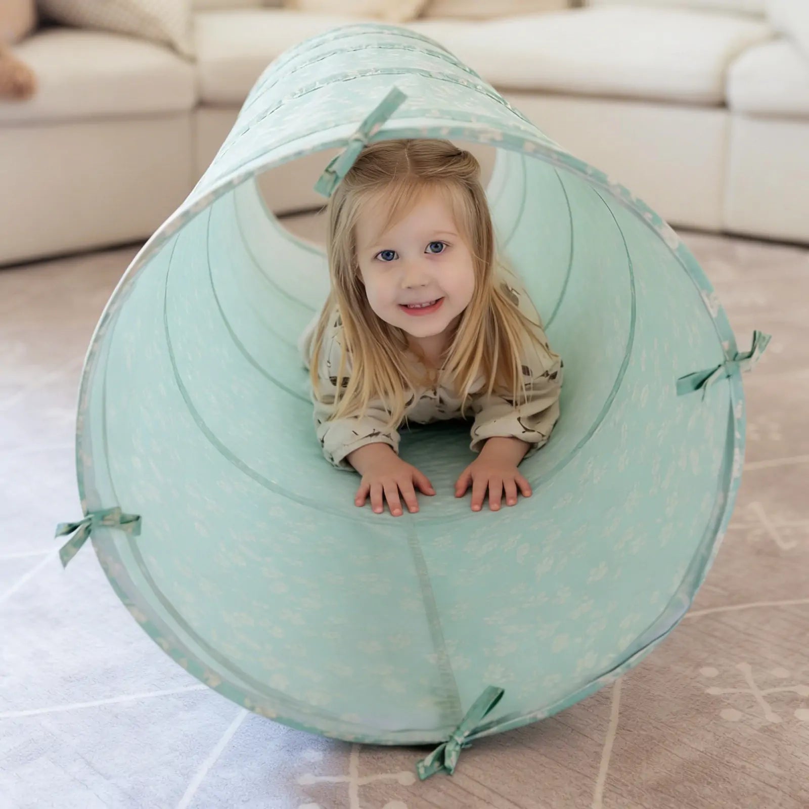 A young girl with blonde hair and a light-patterned shirt smiles at the camera as she crawls through the Tiny Land® Adventure Play Tunnel - Green by Tiny Land, set up on a patterned carpet in the living room.|green