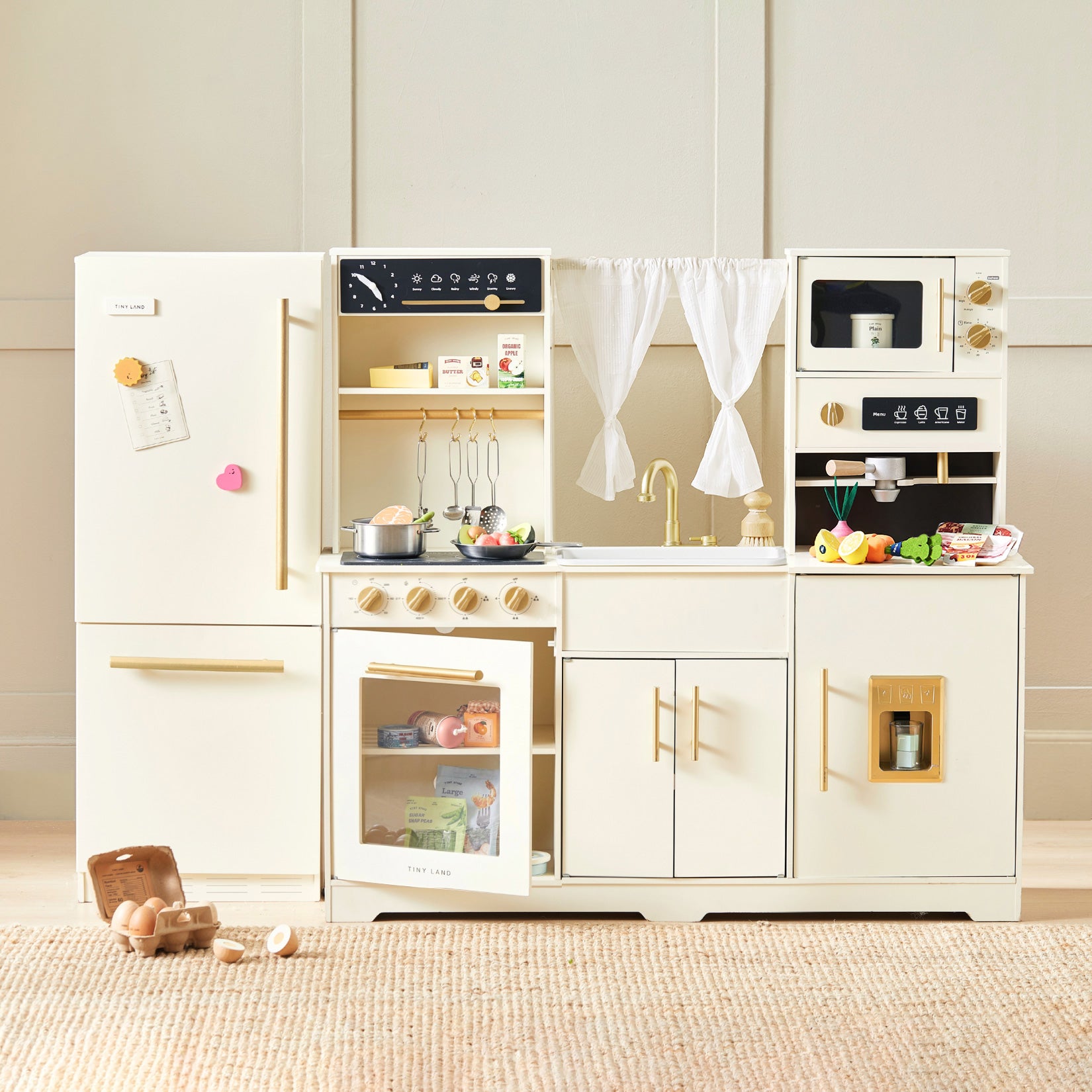 Introducing the Tiny Land® Iconic Water Play Kitchen & Premium Wooden Refrigerator by Tiny Land INC—a cream-colored toy kitchen set equipped with a fridge, oven, stove, sink, and microwave. Styled for endless pretend play, it features realistic play food, utensils, and accessories. The cozy setup is enhanced with a woven rug beneath the set and the charming Premium Wooden Refrigerator that adds to the playful atmosphere.
