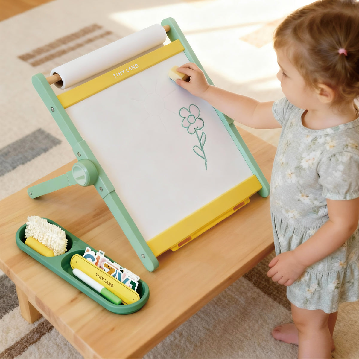 A young child uses the Tiny Land® Double-Sided Magnetic Tabletop Easel - Green, erasing a flower drawing. Art supplies and a tray with markers and cards are on the table next to the Tiny Land easel.