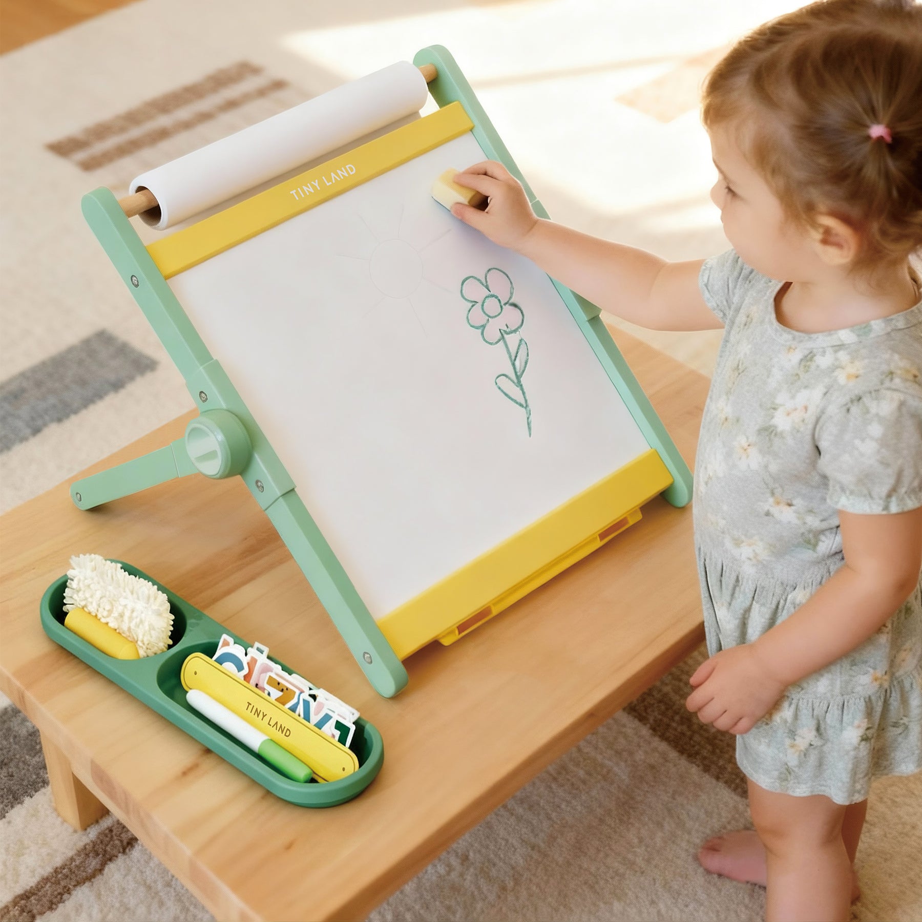 A young child uses the Tiny Land® Double-Sided Magnetic Tabletop Easel - Green, erasing a flower drawing. Art supplies and a tray with markers and cards are on the table next to the Tiny Land easel.