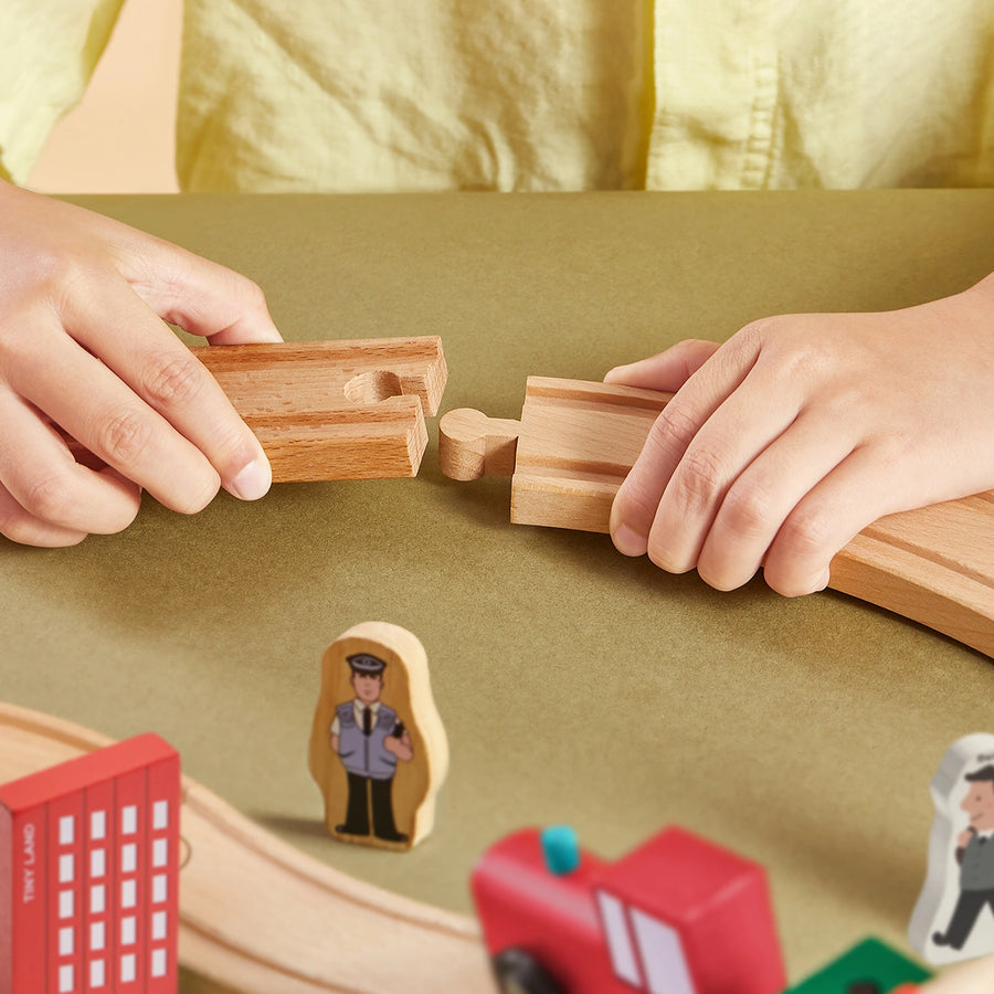 A child connects pieces of the Tiny Land® Wooden Train Set for Children 39 Pcs on a table. Wooden figures—a policeman, a red vehicle, and a red building—add fun, making this Tiny Land toy perfect for early learning.|39 Pcs
