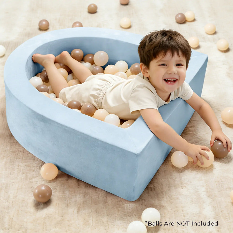 A smiling child relaxes in the Tiny Land® PlayNest Soft Foam Ball Pit by Tiny Land, designed in a blue raindrop shape and filled with beige, white, and brown plastic balls for sensory play. Some balls are scattered across the carpeted floor. Text reads: “*Balls are NOT Included.”.|Blue