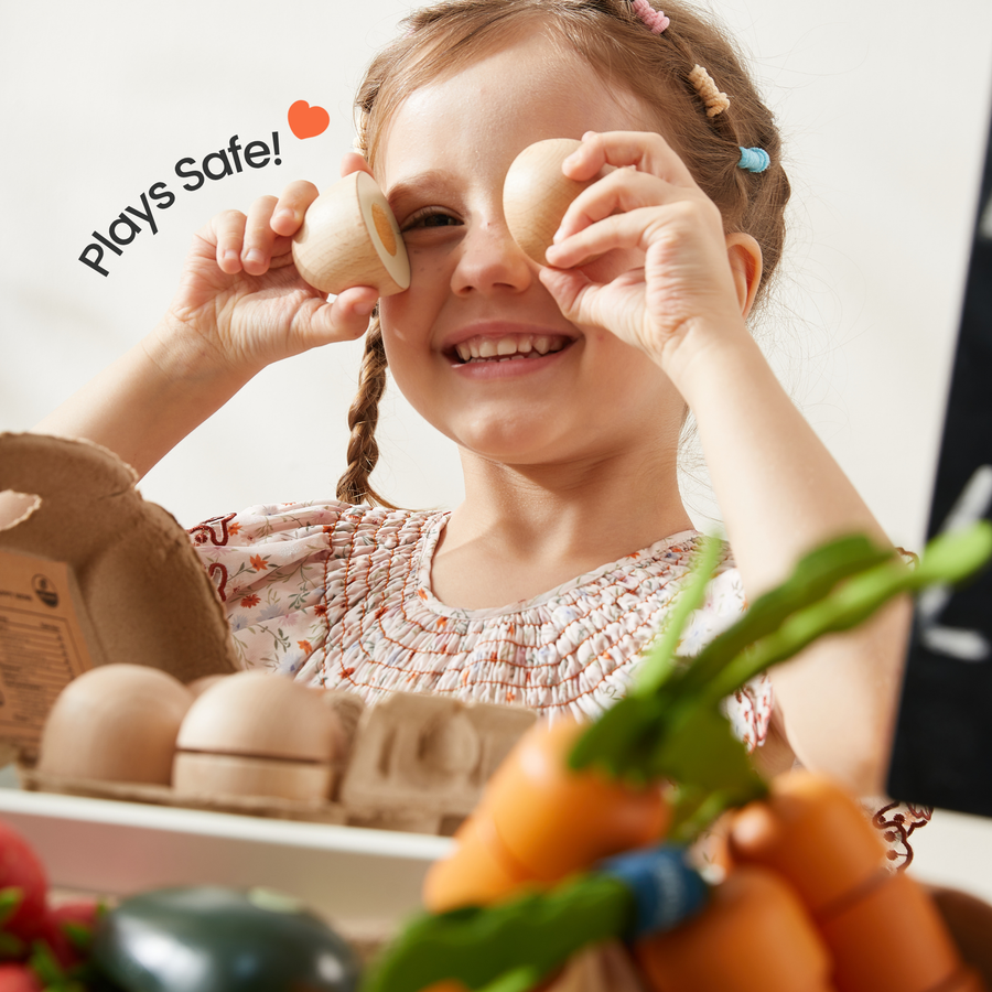 A smiling young girl holds two eggs as glasses. Text reads "Plays Safe!" with a heart. Colorful toy fruits from the Tiny Land® Organic Play Food Collection by Tiny Land Play Food make playtime fun for toddlers in the foreground.