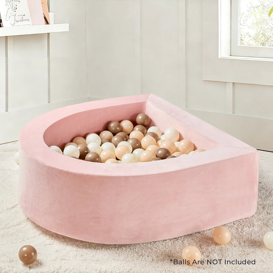 A soft, pink, crescent-shaped Tiny Land® PlayNest Soft Foam Ball Pit—balls not included—sits on a plush rug, perfect for sensory play and ready to be filled with pastel or neutral-colored balls. Beige paneled walls are visible in the background. Text in the corner reads, *Balls Are NOT Included.|Pink