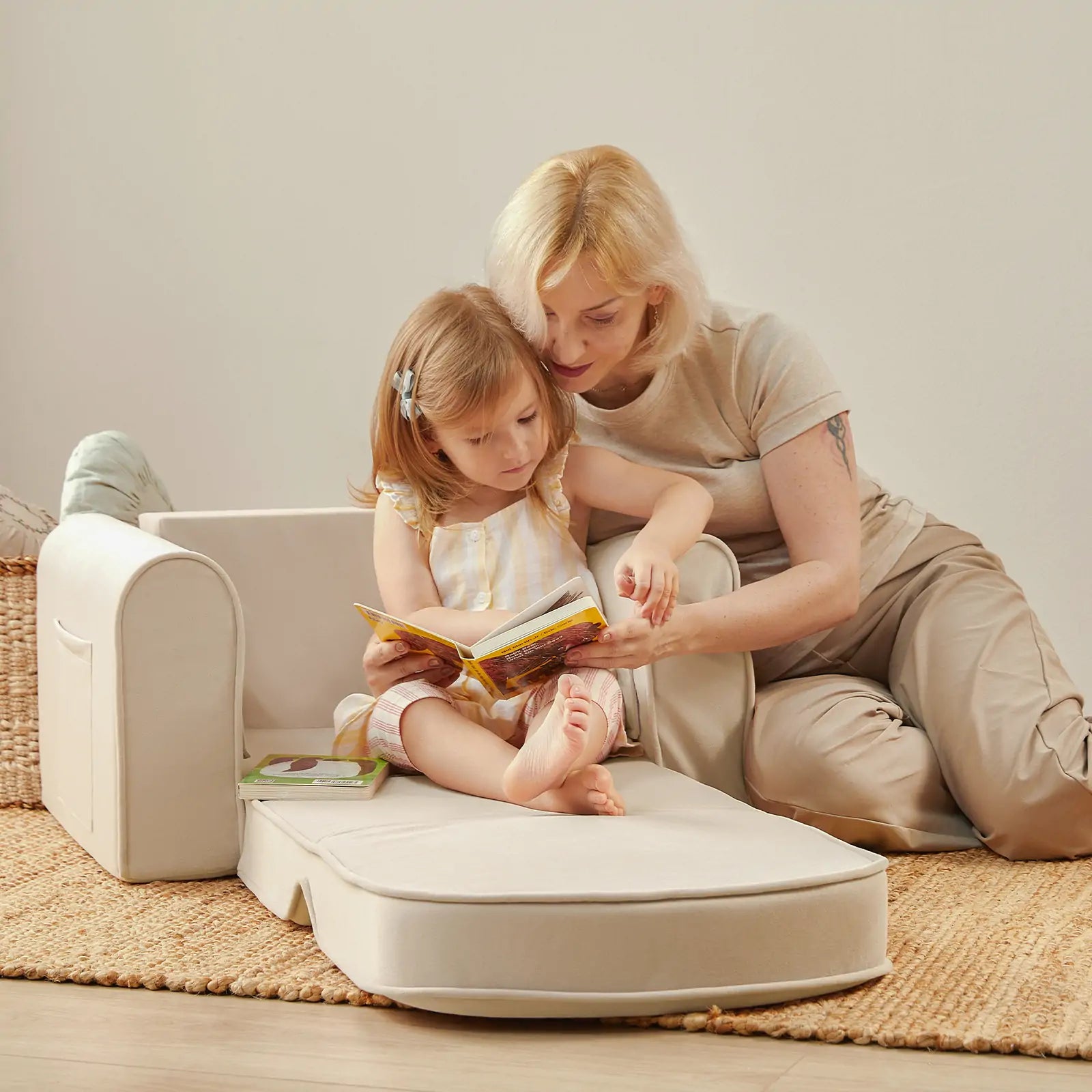 A woman and a young girl sit on the cream-colored Tiny Land® GentleSnug Kid Chair by Tiny Land. The woman helps the girl read a book, both looking focused and happy. The cozy, bright scene features a woven basket in the background.|Beige