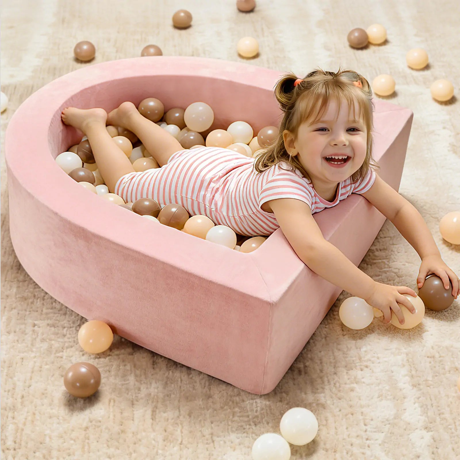 A young girl smiles as she lounges in the Tiny Land® Pink PlayNest Ball Pit by Tiny Land INC, enjoying sensory play in a soft, crescent-shaped pit (Ocean Ball not included) on a light carpeted floor.|pink