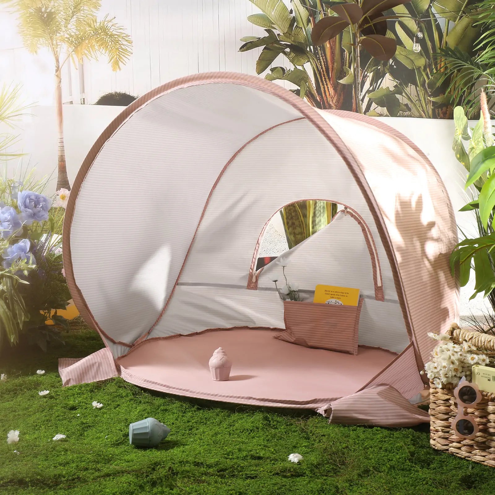 The Tiny Land® One-Stop Beach Toy & Tent Set – Blush Pink, a small UV-protected pink and white striped tent, is set up on grass among flowers, a basket, and plants with sunlight streaming in from the left.