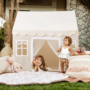 A young girl lies on a circular mat in front of a Tiny Land® Cozy Nook – GentleSnug Chair & Playhouse Tent, while a toddler stands beside her next to a low table with toy drinks.|White
