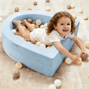 A smiling young child lies in the Tiny Land® PlayNest Ball Pit with 200 Ocean Balls—heart-shaped and blue, filled with white, beige, and brown balls—reaching to play on a beige carpet in this inviting toddler play space by Tiny Land.|Blue