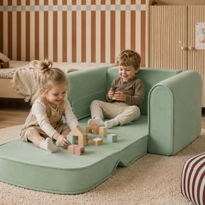 Two young children play with wooden blocks while sitting on the Tiny Land® GentleSnug Kids Sofa Chair by Tiny Land. The cozy playroom has a striped accent wall, beige carpet, and light wood furniture in the background.