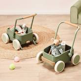 Two Test Tiny Land® JoyRide Baby Walker with Blocks - Sage sit on a round woven rug, each filled with stuffed animals and toys. Pastel balls are scattered nearby on the wooden floor.