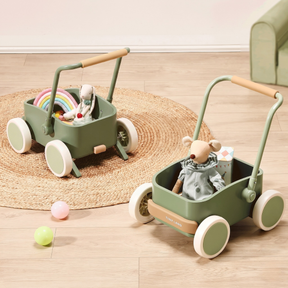 Two Test Tiny Land® JoyRide Baby Walker with Blocks - Sage sit on a round woven rug, each filled with stuffed animals and toys. Pastel balls are scattered nearby on the wooden floor.