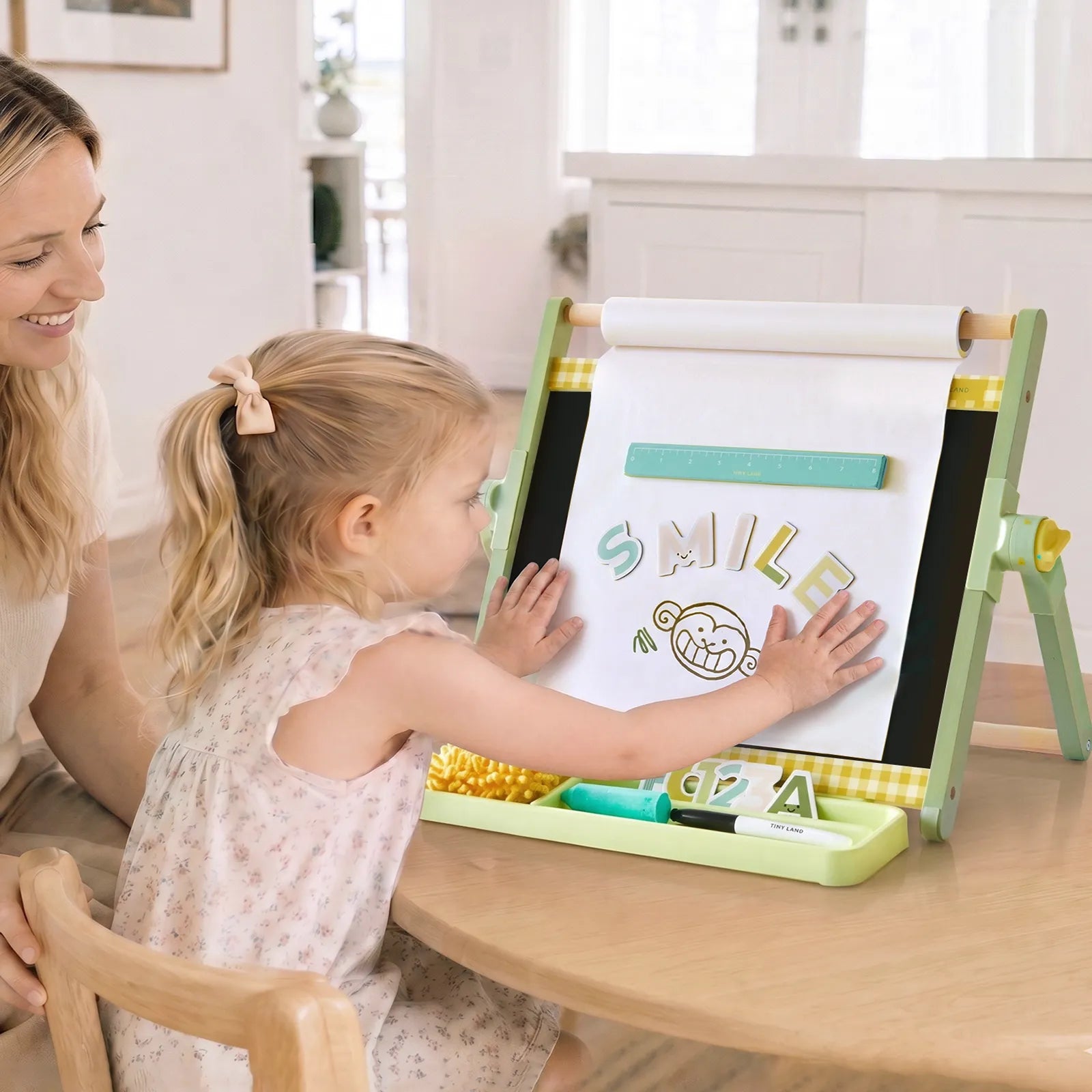 A young girl sits at a table, drawing on the Tiny Land® Double-Sided Magnetic Tabletop Easel while an adult woman smiles beside her. The easel holds a sheet of paper where she has written “SMILE” and drawn a cartoon face. Art supplies are neatly organized in the tray beneath the easel, creating a cheerful kids’ art station from Tiny Land.|Green