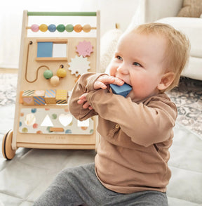 A baby in a brown shirt sits on a mat, chewing on a blue toy block. Behind the baby is the Tiny Land Complete Development Playset — Baby Walker + Activity Cube, featuring colorful beads and shapes that are perfect for encouraging fine motor skills.