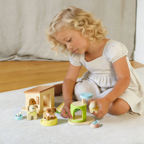 A young child with curly blonde hair and a light dress plays on a white rug with the Test Early Bird Eggs-Tiny Land® Happy Urban Paws pet playset, surrounded by pretend pet toys. A light curtain and wooden floor complete the cozy setting.