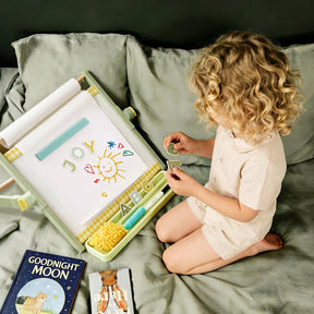 A young child sits on a bed, playing with magnetic letters and drawings on the Tiny Land® Double-Sided Magnetic Tabletop Easel by Tiny Land. The word JOY and a sun are displayed on its magnetic whiteboard. Books, including Goodnight Moon, are nearby.|Green