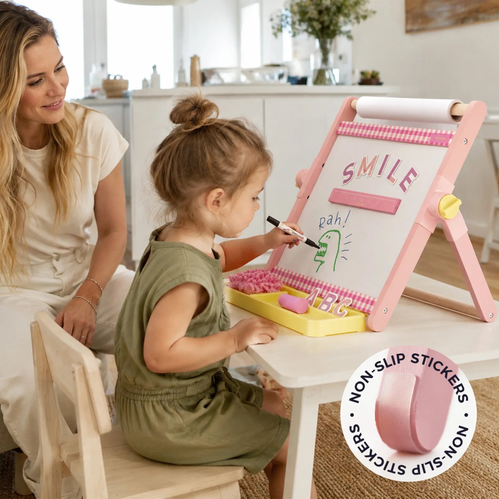 A young girl draws on the Tiny Land® Double-Sided Magnetic Tabletop Easel in pink as a woman smiles nearby in a bright room. A sticker labeled NON-SLIP STICKERS appears in the lower corner.|Pink