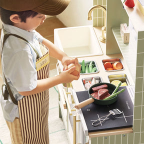 A child in a striped apron plays at the Tiny Land® BistroSwitch Play Kitchen, cracking an egg and using toy veggies and meat on the counter with a pan and utensils, fully immersed in imaginative pretend play by Tiny Land.|Green