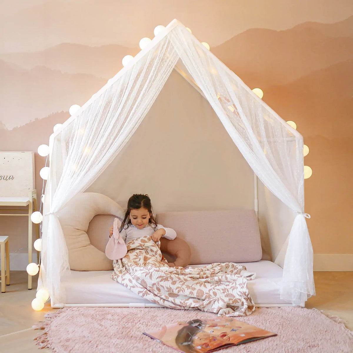 A young girl sits inside the Tiny Land® Chef’s Corner Playset—a cozy cream playhouse and kitchen set—surrounded by soft pillows and a pink rug, with string lights above, reading a book under a patterned blanket.