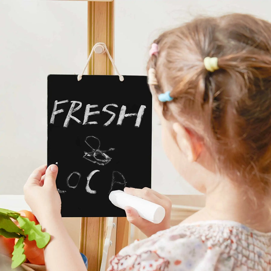 A girl with colorful hair ties writes FRESH LOCAL in white chalk on a blackboard next to her Tiny Land® Kid's Grocery Store Playset, adding fun to pretend shopping as vegetables rest on the wooden table.