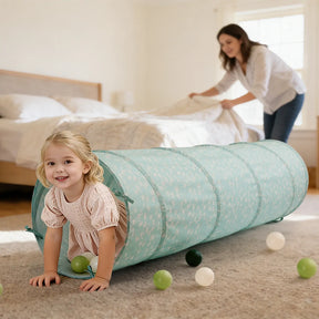 A young girl smiles as she crawls through the Tiny Land® Adventure Play Tunnel - Green, surrounded by plastic balls, while an adult woman makes the bed in a bright bedroom. Brand: Tiny Land.|green