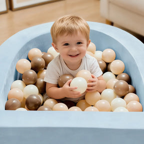 A happy toddler sits in the Tiny Land® Blue PlayNest Ball Pit with 200 ocean balls by Tiny Land INC, holding a ball. The pit is filled with beige, brown, and cream ocean balls. Light furniture and a wooden floor are in the background.|blue