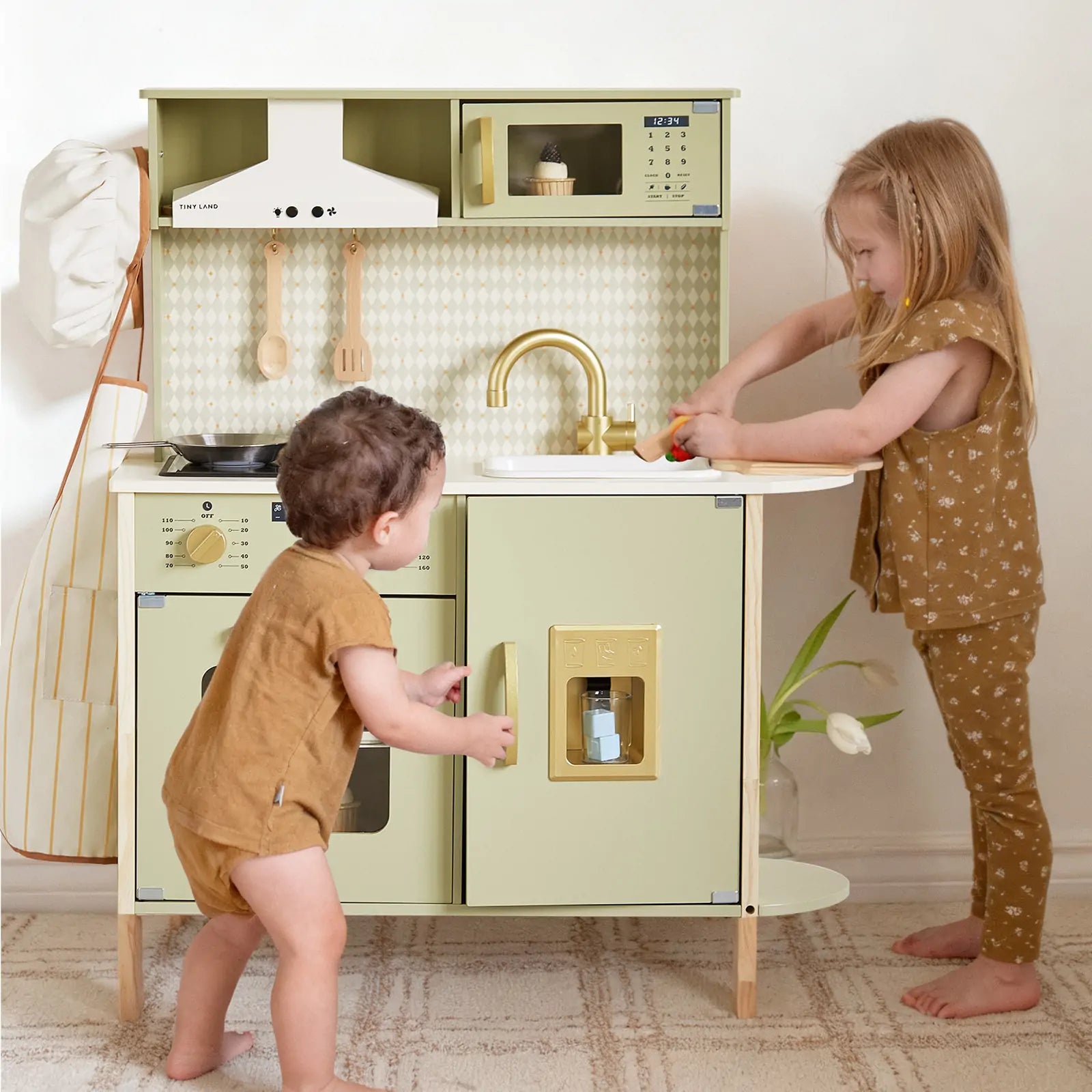 Two young children play with the Tiny Land® PopJoy Play Kitchen - Green. The older child pretends to wash vegetables at the sink, while the younger opens the oven. Both wear brown outfits and stand on a patterned rug. Brand: Tiny Land.