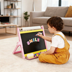 A young child writes with chalk on the Tiny Land® Double-Sided Magnetic Tabletop Easel - Pink, displaying colorful magnetic letters spelling SMILE, while sitting on a carpeted floor in a bright, cozy living room.