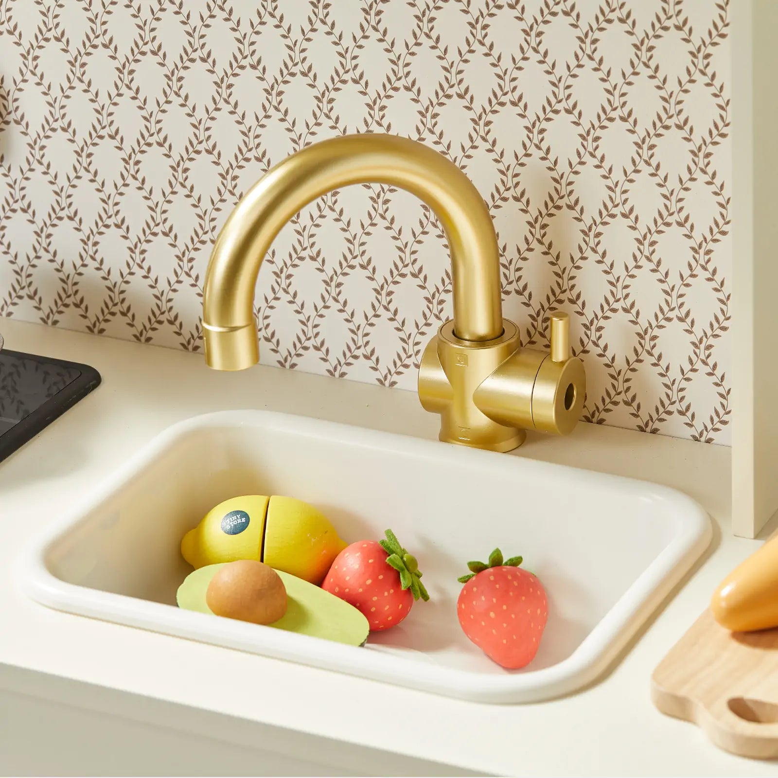 A gold-toned faucet above a white sink with toy food—a sliced avocado, yellow fish, and two strawberries—sparks pretend play in the Tiny Land® PopJoy Kitchen & Fridge Set by Tiny Land, set before wallpaper with small brown leaves.