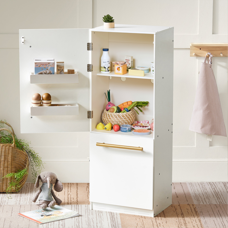 A white toy refrigerator holds the Tiny Land® Organic Fruit & Veggie Pack by Tiny Land, displaying realistic wooden fruits, vegetables, eggs, and drinks inside. A stuffed rabbit sits nearby on the floor. The fridge door is open to reveal neatly arranged pretend groceries. In the background, a basket and an apron hang on display.