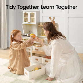 A woman and a young girl tidy up play food in the Tiny Land® Wooden Play Kitchen with Storage for Creative Toddlers. Text above reads, "Tidy Together, Learn Together." Both are dressed in light-colored clothing and kneel on a rug as they carefully organize pretend cookware in their Tiny Land kitchen set.|cream