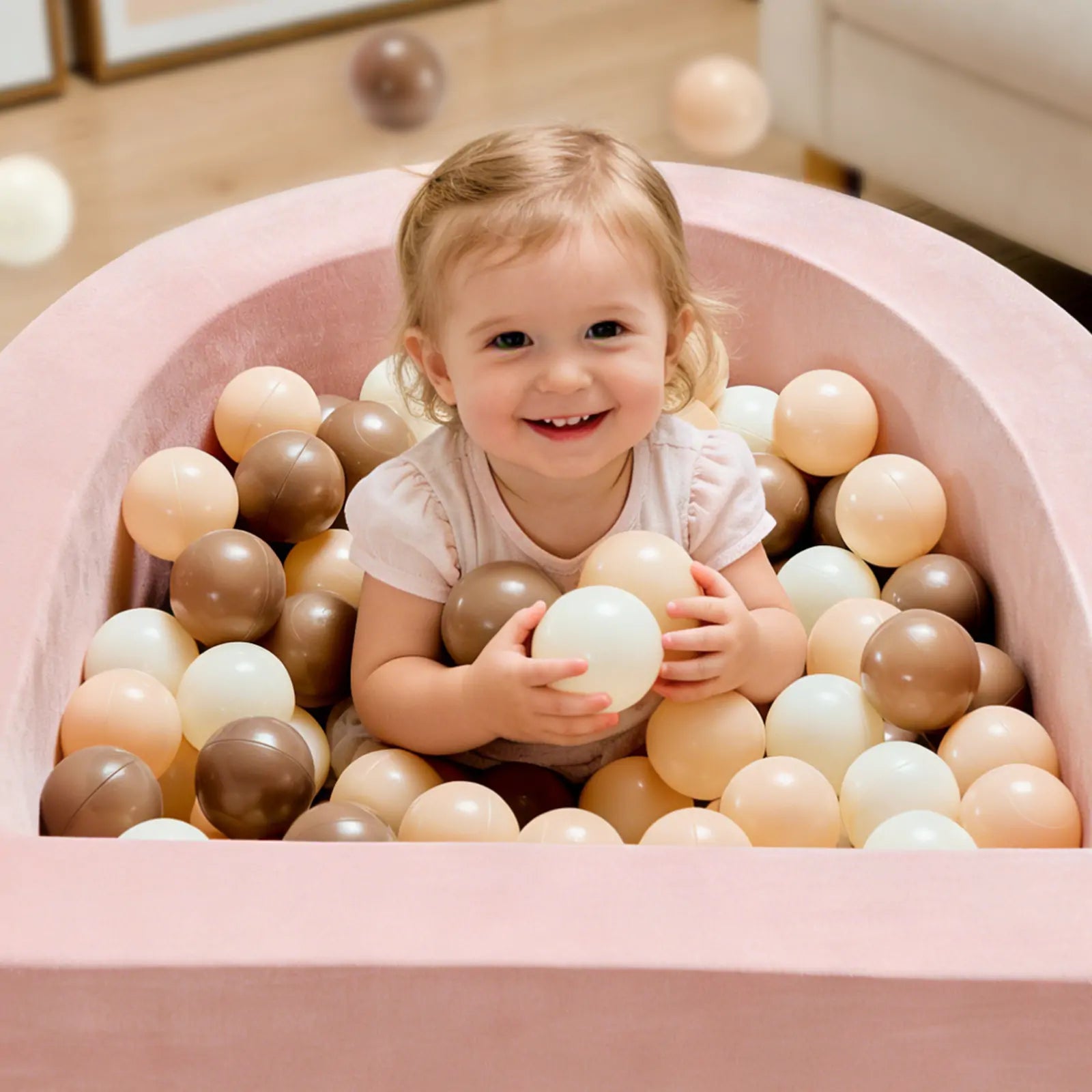 A happy toddler enjoys the Early Bird Eggs-Tiny Land® PlayNest Ball Pit with 200 ocean balls by Tiny Land INC, sitting in a pink foam nest and holding a cream ball among beige, brown, and white balls.|pink