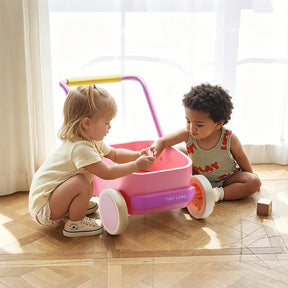 Two young children play together on a wooden floor beside the Tiny Land® 3-in-1 Wooden Baby Walker Wagon in Pink, with sunlight streaming through sheer curtains as one child hands an object to the other.