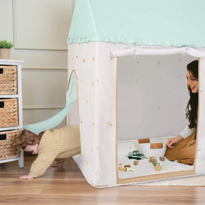 A child crawls out of the back flap of a Tiny Land® Sweet Woodland Kids Play Tent by Tiny Land INC, while an adult smiles inside on a play mat with wooden toys and blocks scattered nearby on a wooden floor beside a wicker-drawer cabinet.