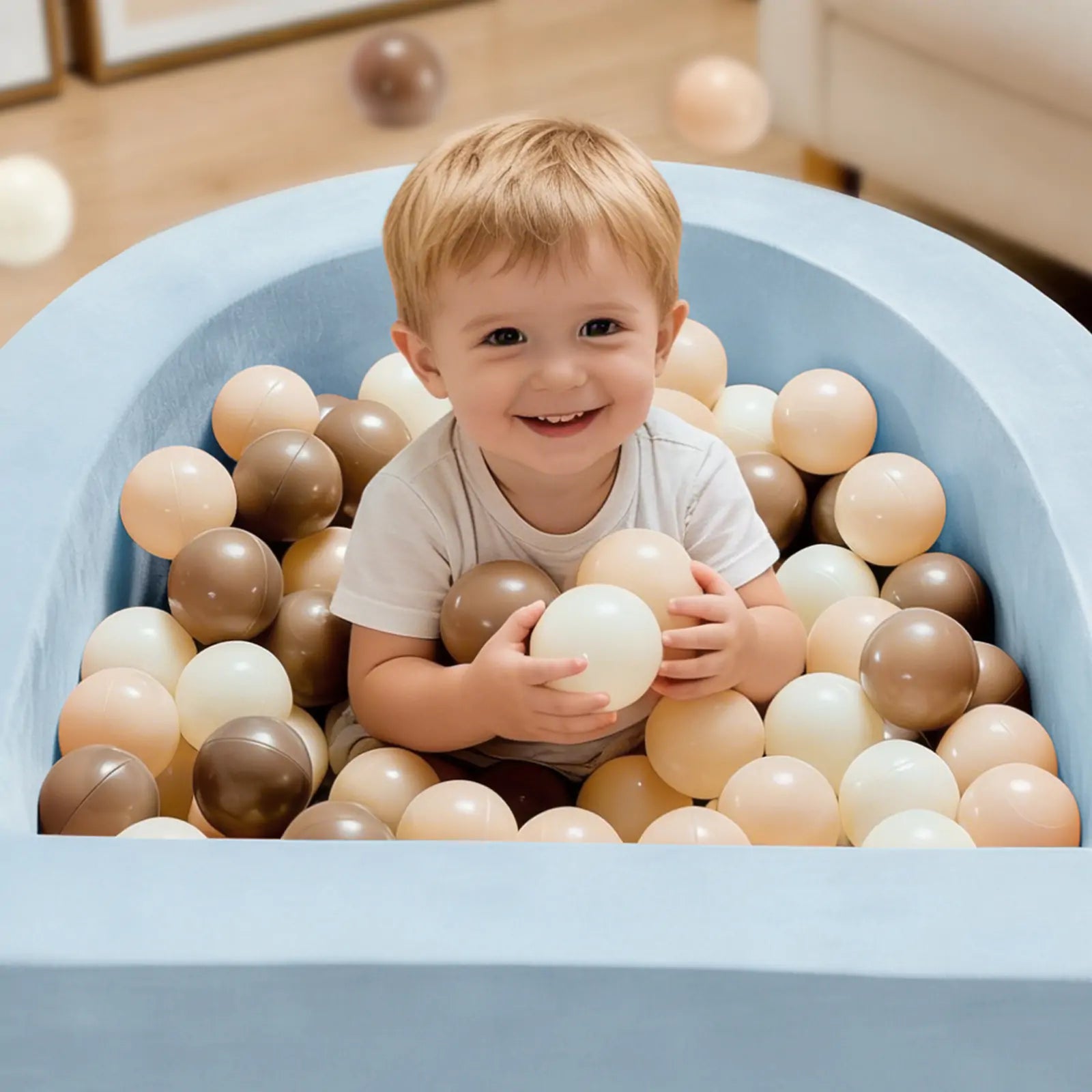 A smiling toddler sits in the Tiny Land® PlayNest Ball Pit with 200 Ocean Balls by Tiny Land, surrounded by blue foam and a mix of beige, brown, and white ocean balls, holding some in their hands.|Blue