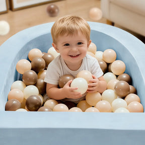 A smiling toddler sits in the Tiny Land® PlayNest Ball Pit with 200 Ocean Balls by Tiny Land, surrounded by blue foam and a mix of beige, brown, and white ocean balls, holding some in their hands.|Blue