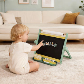 A toddler sits on a carpeted floor, pointing at the Tiny Land® Double-Sided Magnetic Tabletop Easel - Green by Tiny Land, with “SMILE” spelled in white letters. The cozy room includes a sofa, cushions, and soft neutral decor.