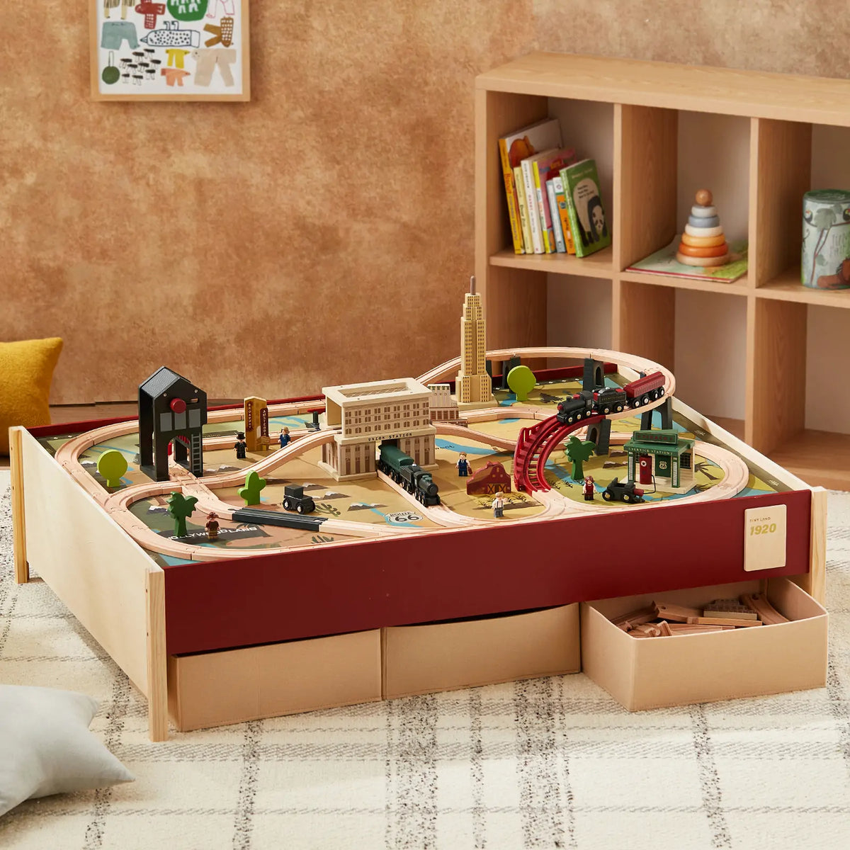 The Tiny Land® 1920 Railway Adventure Train Table by Tiny Land INC features trains, tracks, buildings, and trees on a play table with drawers. The room includes a shelf of books and toys plus a beige carpet with a pillow nearby.