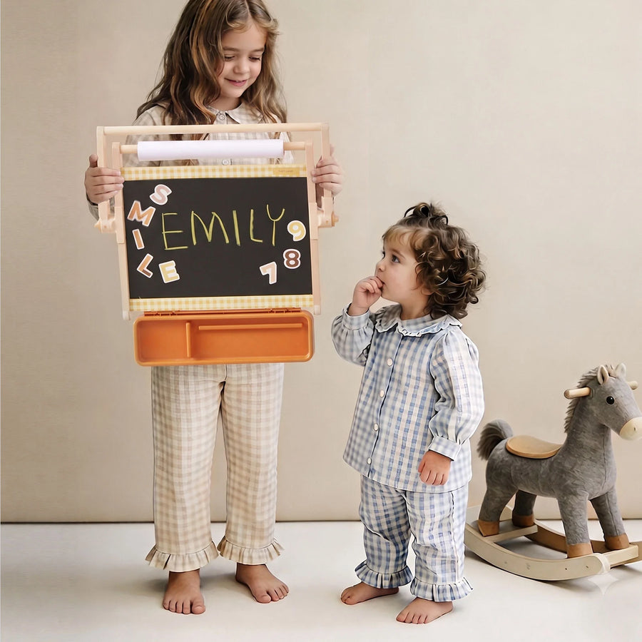 Two young children in pajamas stand barefoot; one holds the Tiny Land® Double-Sided Magnetic Tabletop Easel with “SMILE EMILY 9 18” spelled out in colorful letters. A toy rocking horse is beside them, completing this charming Tiny Land kids art station.|Oatmeal