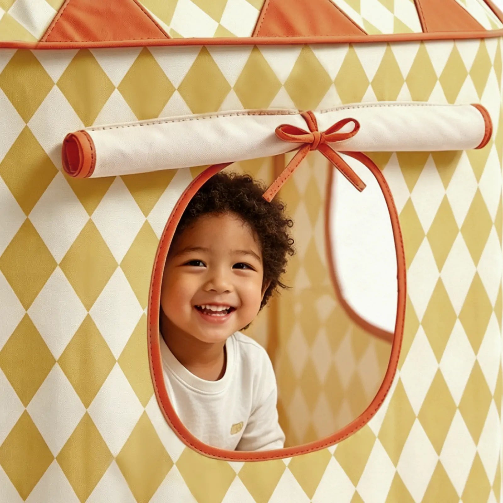 A smiling child with curly hair looks out from the window of the Tiny Land® Tiny Sunshine Castle Tent, featuring a yellow and white diamond pattern with orange trim.