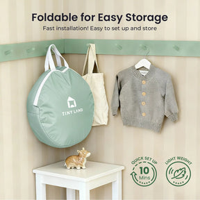 A sage green Tiny Land® Compact Adventure 3-in-1 Play Tunnel (no balls) hangs on a wall hook next to a child’s gray sweater, with a decorative hippo below. Lightweight and easy to set up—perfect for storing your play tunnel.