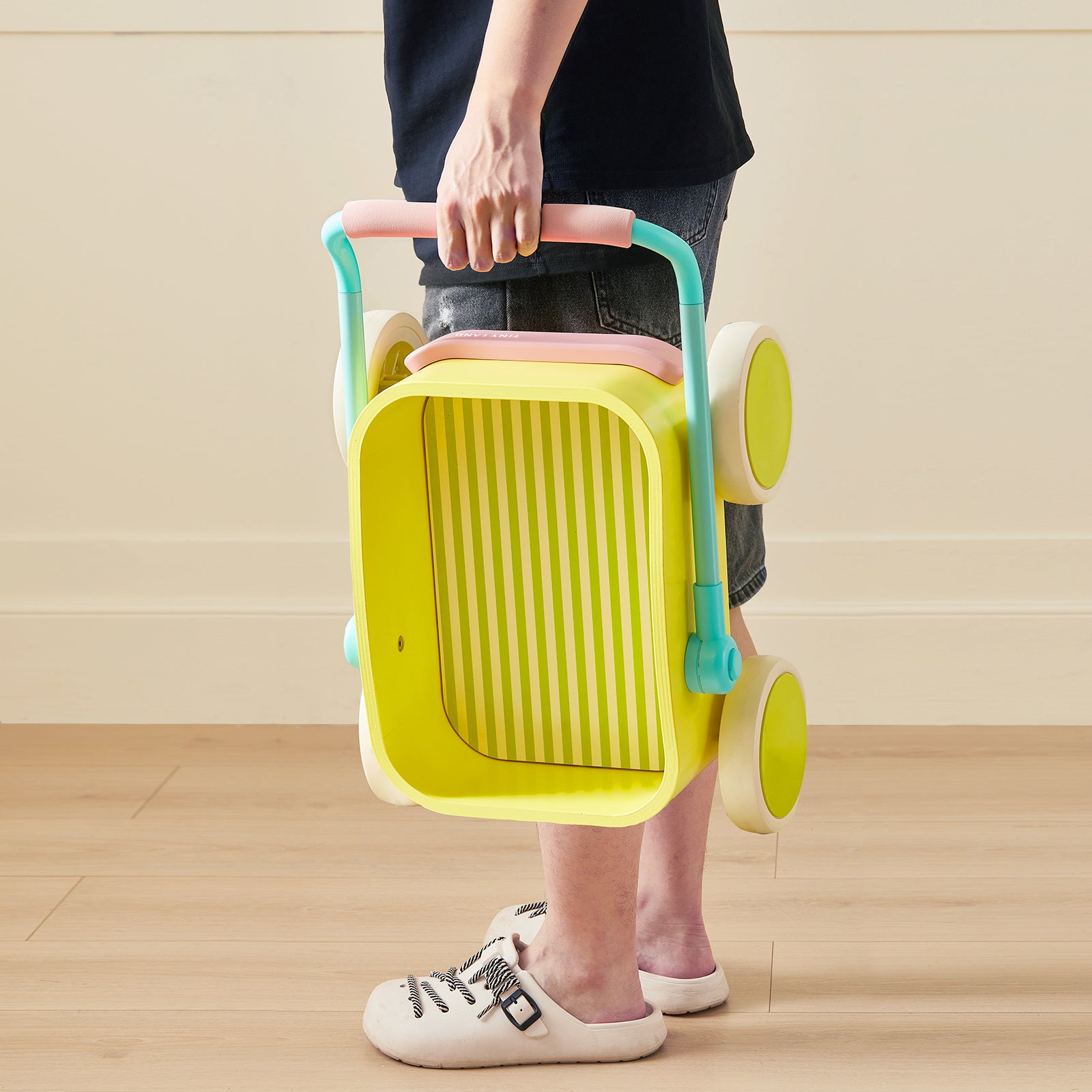 A person in shorts and sneakers holds the Test Tiny Land® JoyRide Baby Walker with Blocks - Spring Color, featuring a yellow striped interior and large white wheels, standing on a wooden floor.
