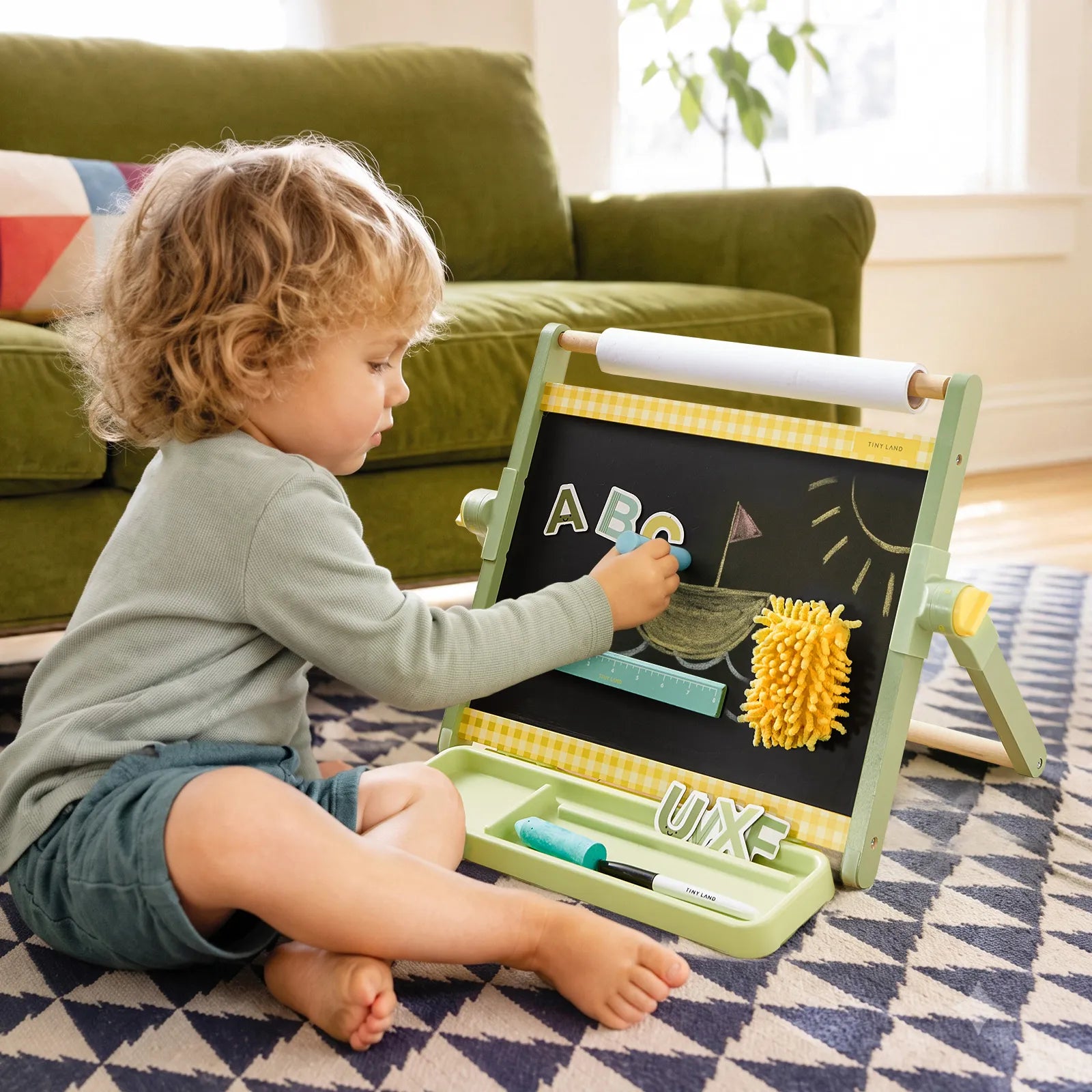 A young child sits on a patterned rug, drawing with colorful letters and chalk on the Tiny Land® Double-Sided Magnetic Tabletop Easel by Tiny Land, in a cozy living room with a green sofa in the background.|Green
