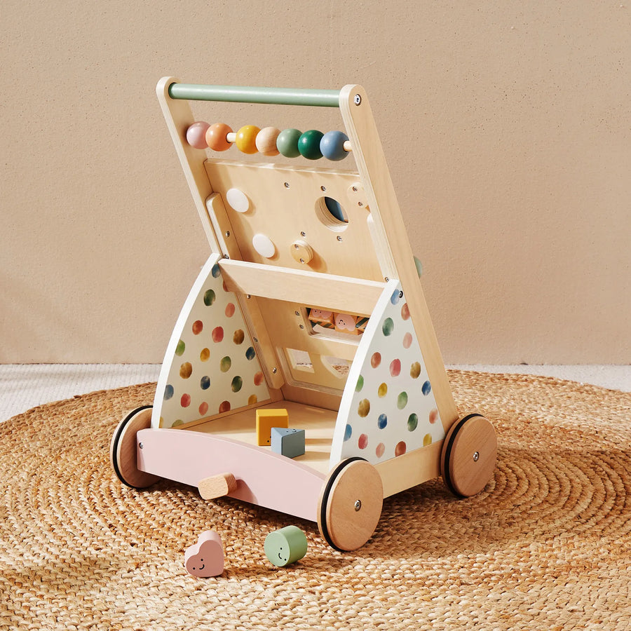 The Tiny Land® Versatile Natural Wooden Baby Walker by Tiny Land, featuring colorful beads, shape sorter holes, and multi-colored polka dots on its sides, is placed on a woven rug with several wooden shapes scattered nearby.