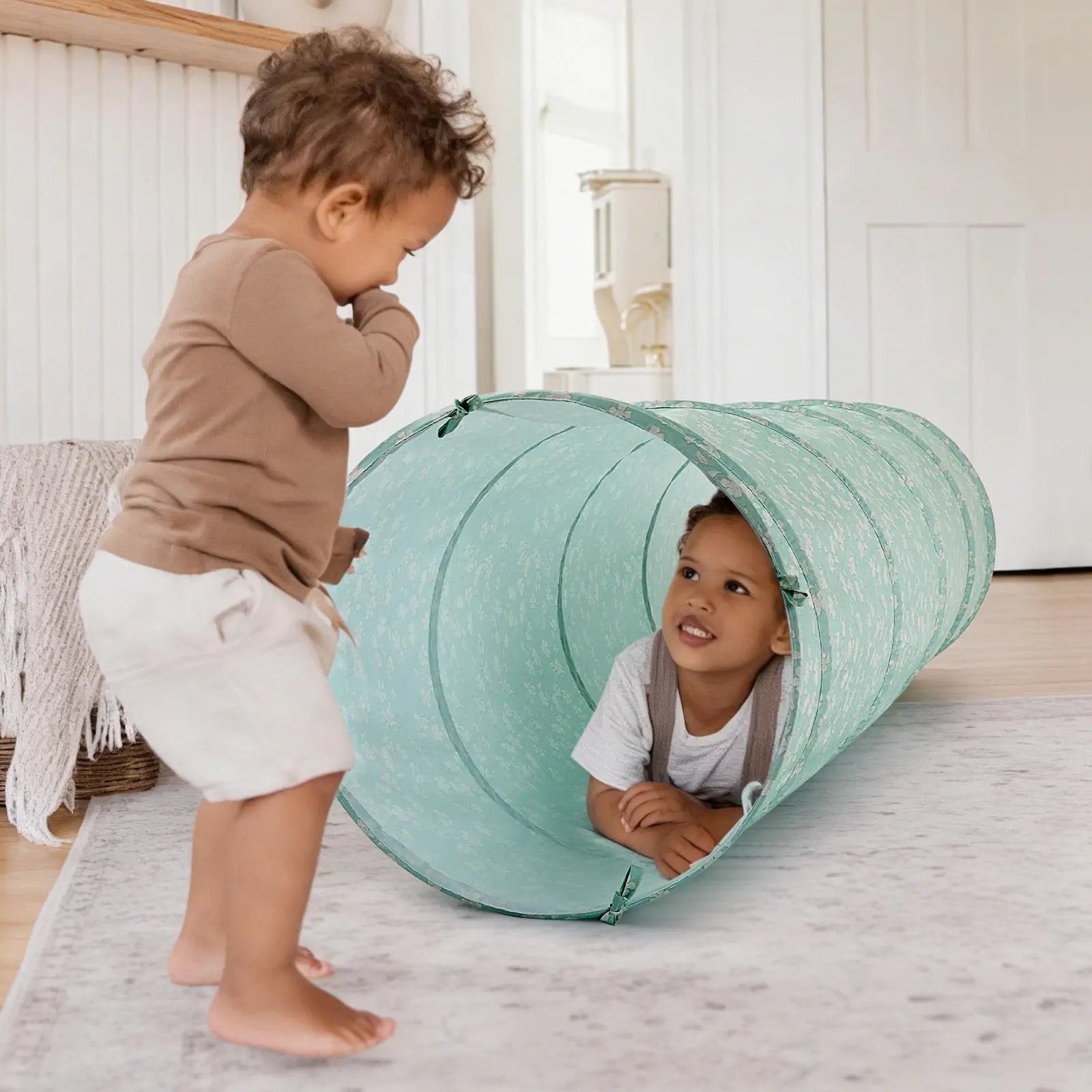 Two young children play indoors; one stands on a rug while the other smiles and looks up at their friend, crawling through the Tiny Land® Adventure Play Tunnel - Green by Tiny Land.|green