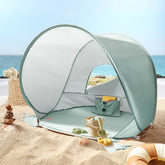 The Tiny Land® One-Stop Beach Toy & Tent Set – Sea Blue provides UV-protective shade on the sand, with included digging tools and a wicker basket nearby, all set against a serene sea and blue sky.