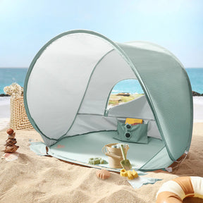 The Tiny Land® One-Stop Beach Toy & Tent Set – Sea Blue provides UV-protective shade on the sand, with included digging tools and a wicker basket nearby, all set against a serene sea and blue sky.